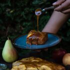 Easy, No Mixer Apple Cake with Salted Caramel Sauce www.thefoodiecorner.gr Photo description: A plate with a piece of cake on it being held above a small table outdoors. A hand is pouring caramel over the cake from a small spoon. On the table below is the cake, some apples, a pear and a knife. In the background is some dense foliage.