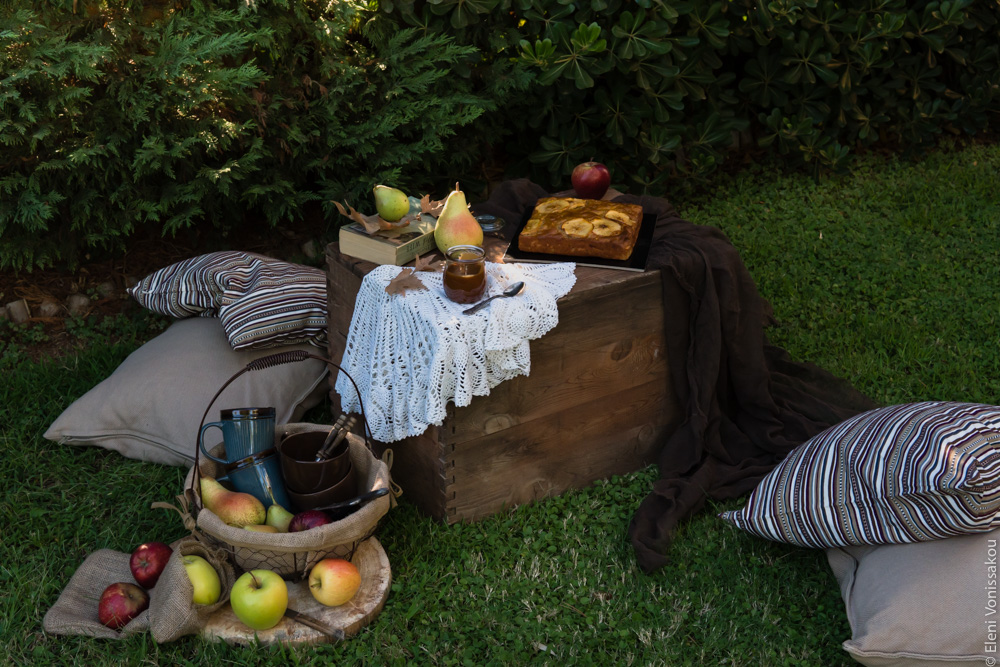 Easy, No Mixer Apple Cake with Salted Caramel Sauce www.thefoodiecorner.gr Photo description: A view from further away of the whole picnic setup. Cusions are lying on the lawn around the small wooden box that is acting as a table. The basket with the mugs is next to it, surrounded by some apples. On the table are the cake, the caramel and a book.