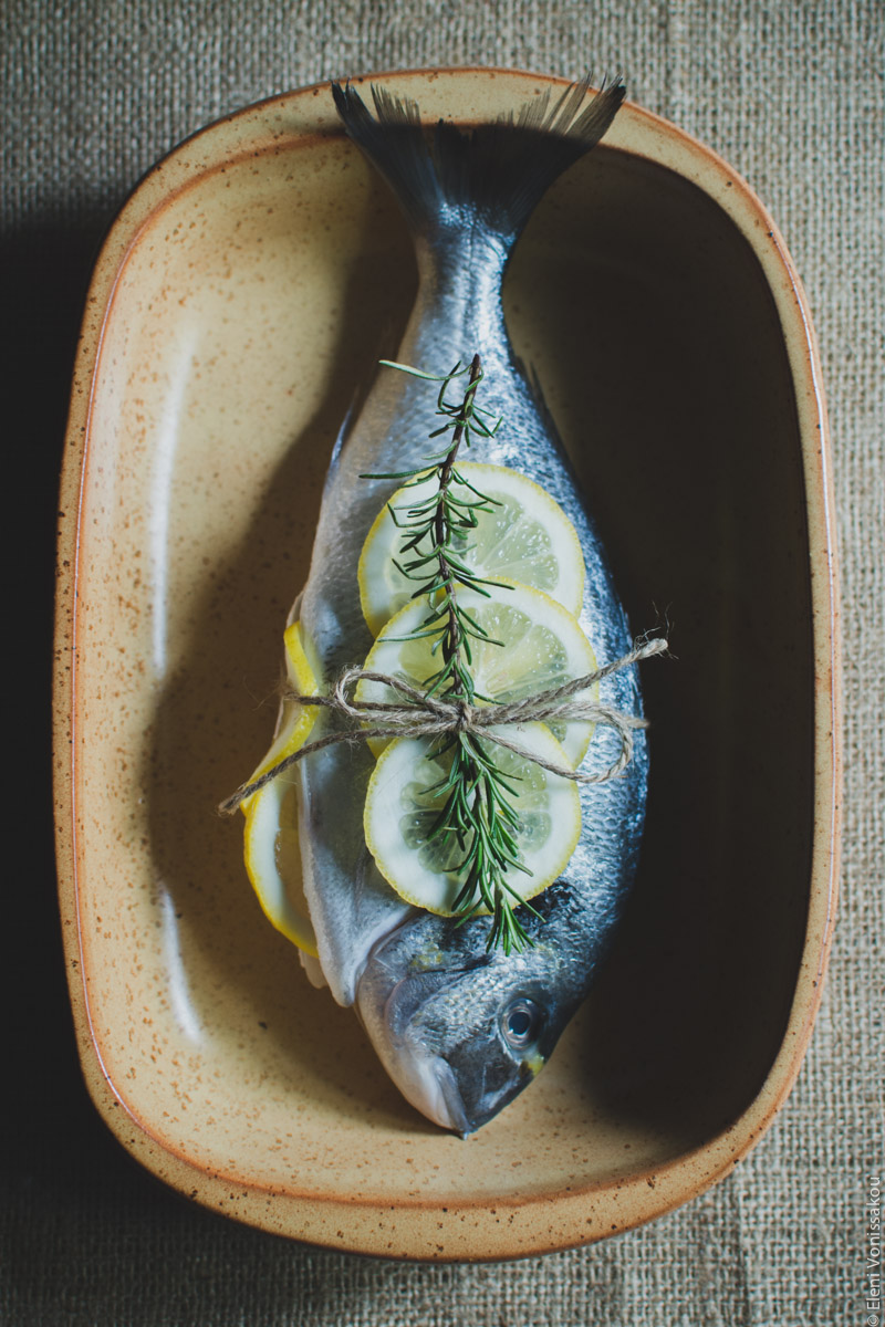 Capturing Athens Food Styling and Photography Workshop www.thefoodiecorner.gr Photo description: A raw fish lying on a baking dish. There are a couple of lemon slices and a sprig of rosemary on top, and some string tied around its belly. some lemon is just visible from inside the cavity..