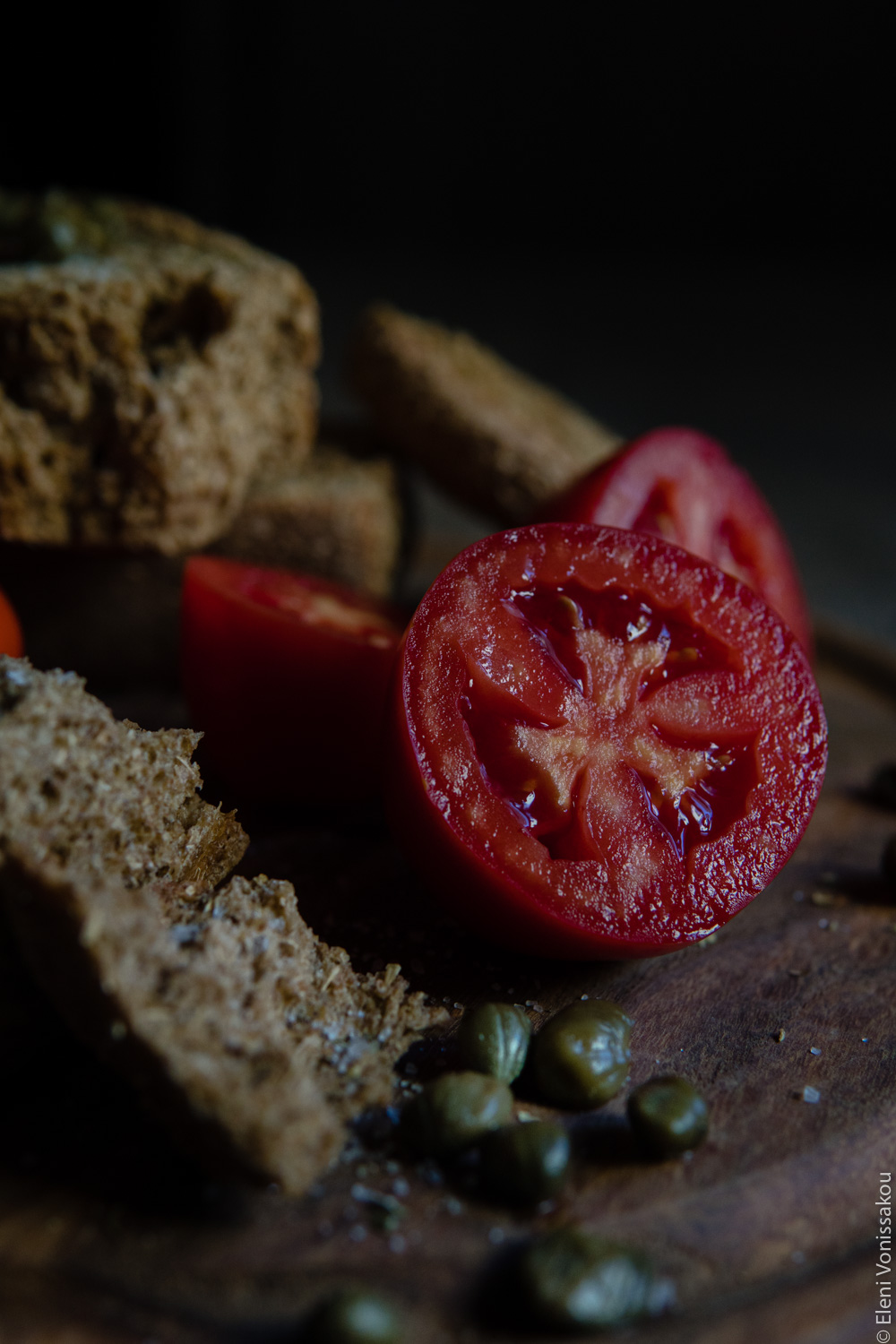 Baked Potato Dakos Salad, with Barley Rusks, Tomato and Feta www.thefoodiecorner.gr Photo description: A close view of a halved tomato sitting on a wooden chopping board, among some pieces of barley rusk. Some capers are also scattered around it.