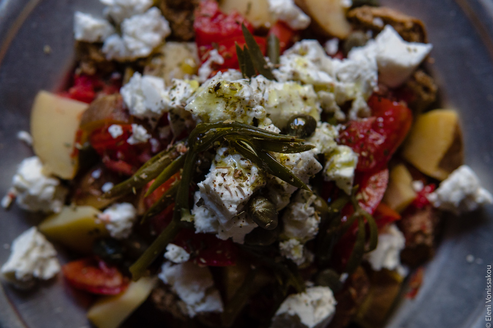 Baked Potato Dakos Salad, with Barley Rusks, Tomato and Feta www.thefoodiecorner.gr Photo description: A very close view of the prepared salad with droplets of olive oil visible on the crumbled feta. Under that are the pieces of potato, tomato and rusks, and on top of the feta are scattered pieces of sea fennel, some capers and some oregano.