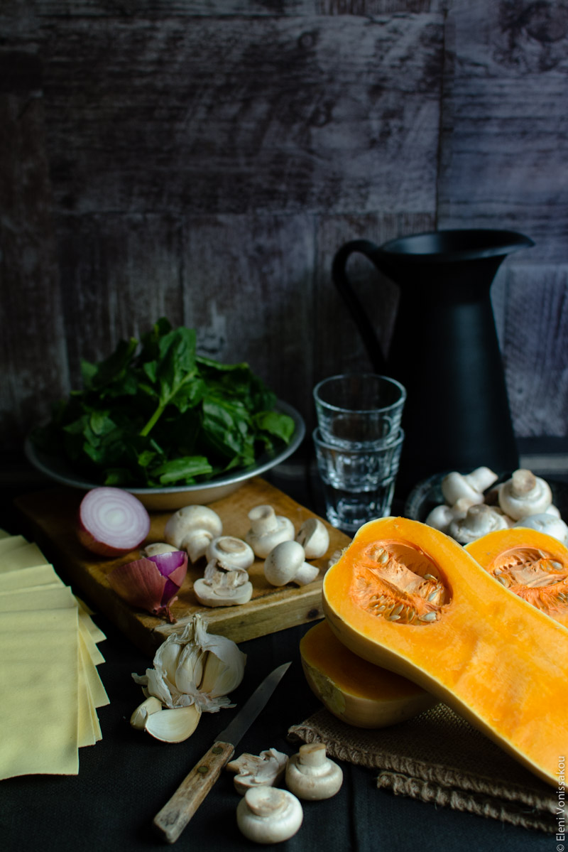 Cottage Cheese Vegetable Lasagne with Butternut Squash, Spinach and Mushrooms www.thefoodiecorner.gr Photo description: An ingredient shot, with the main stars of the dish laid out on a dark surface. A bowl of chopped spinach is in the background, some empty wine glasses next to it and a small bowl of raw mushrooms is to their right, in front of a black jug. To the right of the photo is a cut butternut squash lying face up. In the centre is a chopping board with a halved onion and chopped mushrooms on it. To the left are some sheets of lasagne and a broken up head of garlic. To the front are some more mushrooms and a small knife with a wooden handle.