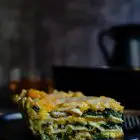Cottage Cheese Vegetable Lasagne with Butternut Squash, Spinach and Mushrooms www.thefoodiecorner.gr Photo description: Side view of a piece of lasagna on a black plate, with visible layers of lasagna pasta, spinach with mushrooms peeking out, and butternut squash puree. The plate is sitting on a piece of burlap and there is a black jug visible in the dark background. The small glasses of wine can be seen just behind the lasagna.
