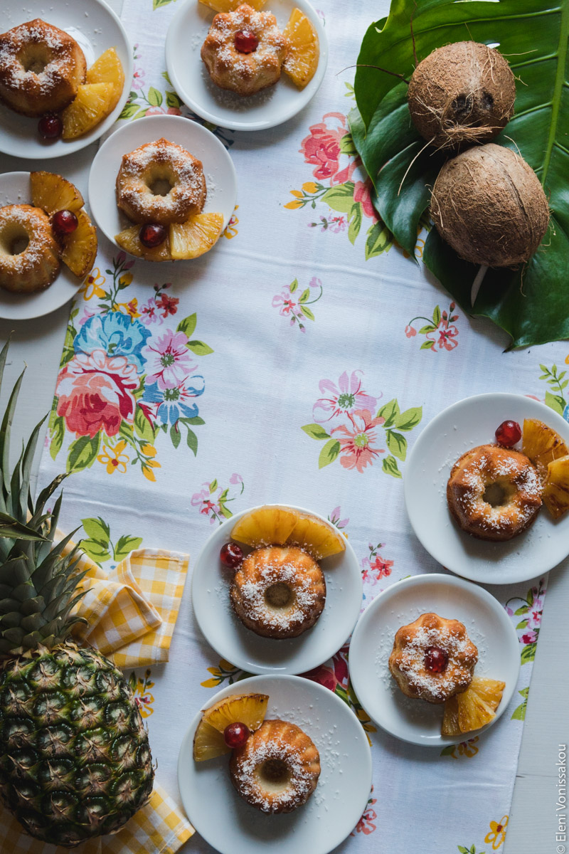 "Ραβανί" με Άρωμα Pina Colada www.thefoodiecorner.gr Photo description: Mini bunt shaped ravani cakes on small white plates sitting on a white surface. Some of them are on a narrow white tablecloth with colourful flowers on it. The plates are mainly in the top left and bottom right corners. Top right are two coconuts on a big leaf and bottom left is a pineapple on a bright coloured tea towel.