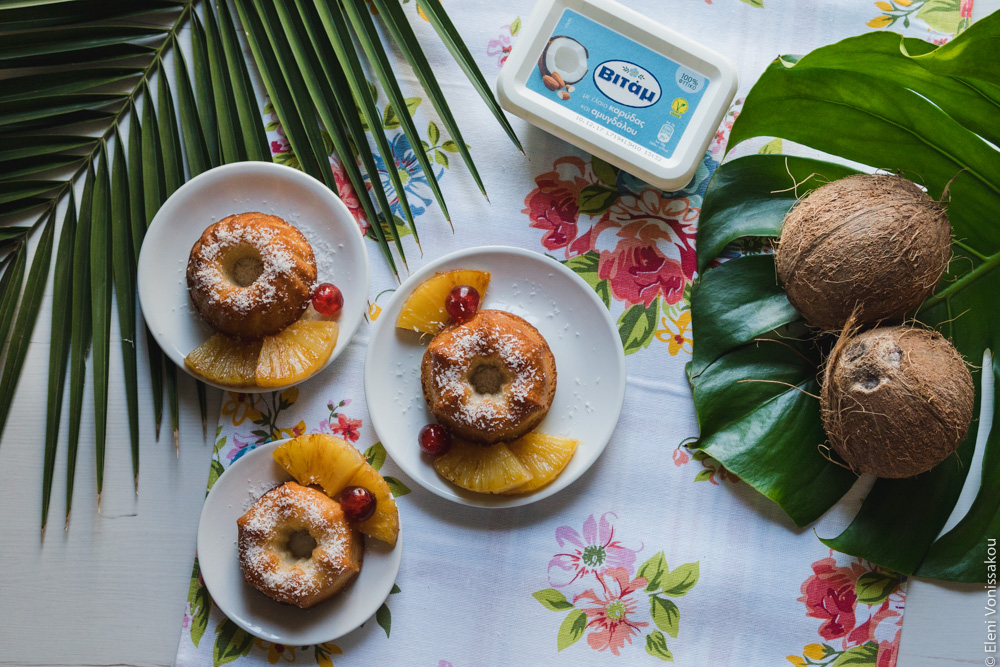 "Ραβανί" με Άρωμα Pina Colada www.thefoodiecorner.gr Photo description: Three mini bunt shaped cakes on small plates sitting close together on a flowery tablecloth. To the top left is a large spiky palm tree branch, and to the right a large monstera leaf with two coconuts sitting on it. Top centre is a tub of coconut flavoured Vitam margarine.