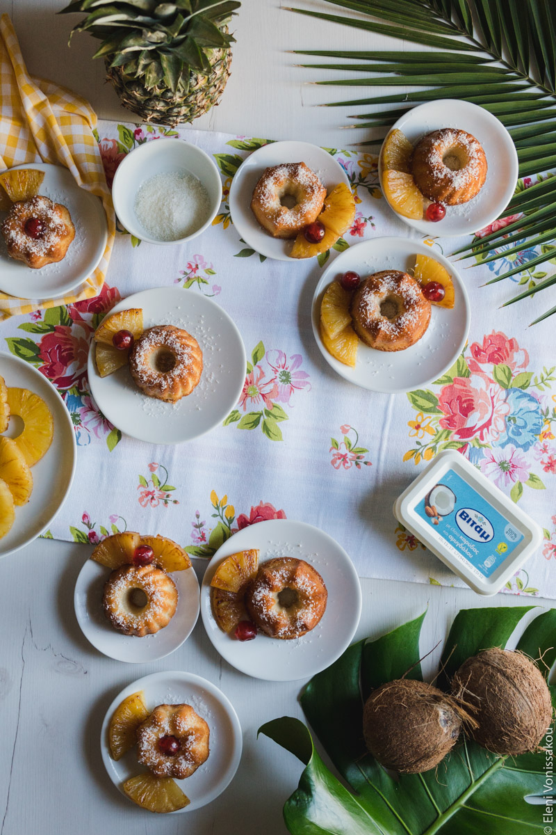 "Ραβανί" με Άρωμα Pina Colada www.thefoodiecorner.gr Photo description: Lots of cakes all on a flowery tablecloth. The corners of the photo are adorned with leaves, a pineapple and coconuts. There is also a plate of pineapple slices half visible on the left side, and a bowl of coconut flakes.