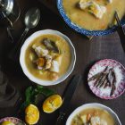 Orange and Lemon Fish Soup with Cod. My version of Greek Psarosoupa. www.thefoodiecorner.gr Photo description: Top view of two bowls of soup, plus one larger serving bowl with a soup ladle in it. The soup is thick and there are vegetables and pieces of fish protruding from within. To the right is a small plate with anchovies, to the bottom left a small plate with two halves of a lemon. There are also pieces of orange, slightly squeezed, near the plate of lemons, and a knife with a wooden handle. Scattered around are some celery leaves. Towards the top left are a couple of soup spoons and a metal olive oil jug, and at the top in the centre are some more orange pieces on a wooden chopping board. Everything is on a dark wooden surface.