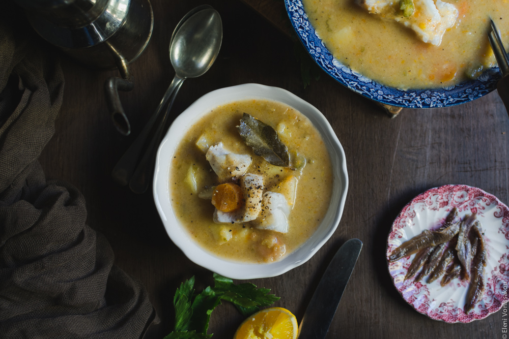 #10 Orange and Lemon Fish Soup with Cod. My version of Greek Psarosoupa. www.thefoodiecorner.gr Photo description: One bowl of soup in the centre of the photo. To the top right (half visible) is the serving bowl. The the top left is the olive oil canister and to the left a brown piece of muslin fabric. To the left the small plate of anchovies.