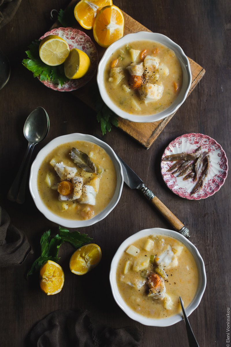 Orange and Lemon Fish Soup with Cod. My version of Greek Psarosoupa. www.thefoodiecorner.gr Photo description: Three bowls of the soup all layed out on a wooden surface. One of them is sitting on a wooden chopping board next to some pieces of orange and lemon. A small plate of anchovies sits to the right.