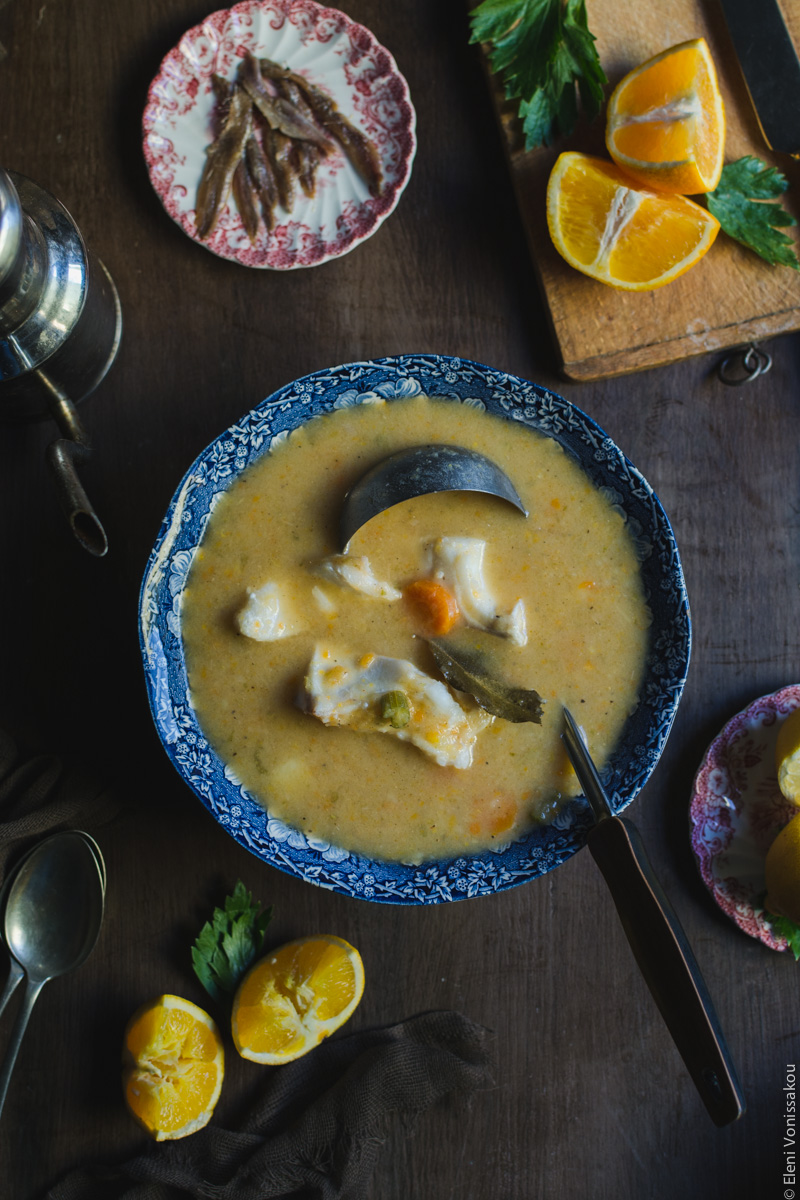 Orange and Lemon Fish Soup with Cod. My version of Greek Psarosoupa. www.thefoodiecorner.gr Photo description: Top view of a large soup serving bowl with a ladle in it. Pieces of vegetables and fish are protruding from within the soup. To the top a small plate of anchovies and a chopping board with pieces of orange on it. To the bottom left two pieces of orange that have been squeezed.