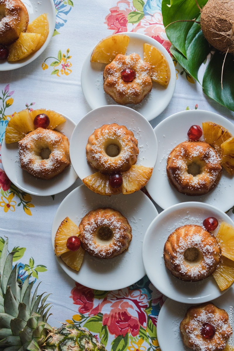 Greek Ravani Turned Mini Pina Colada Cakes www.thefoodiecorner.gr Photo description: Lots of mini bunt shaped cakes on small plates, all placed close to each other, some of them stacked on others. The plates are all in the centre of the photo, sitting on a flowery tablecloth. To the bottom left is a pineapple, only partly visible, and in the top right is a coconut on a green leaf, again barely visible.