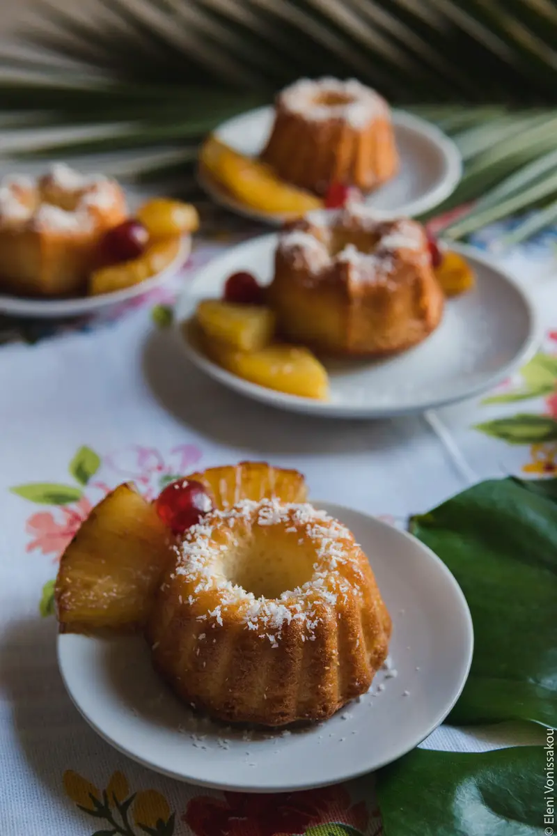 Greek Ravani Turned Mini Pina Colada Cakes www.thefoodiecorner.gr Photo description: A closer view of one of the cakes. Another three cakes are in the background, slightly blurry. The cake has a sprinkling of coconut flakes on it, and some pineapple pieces and a cherry on the side.