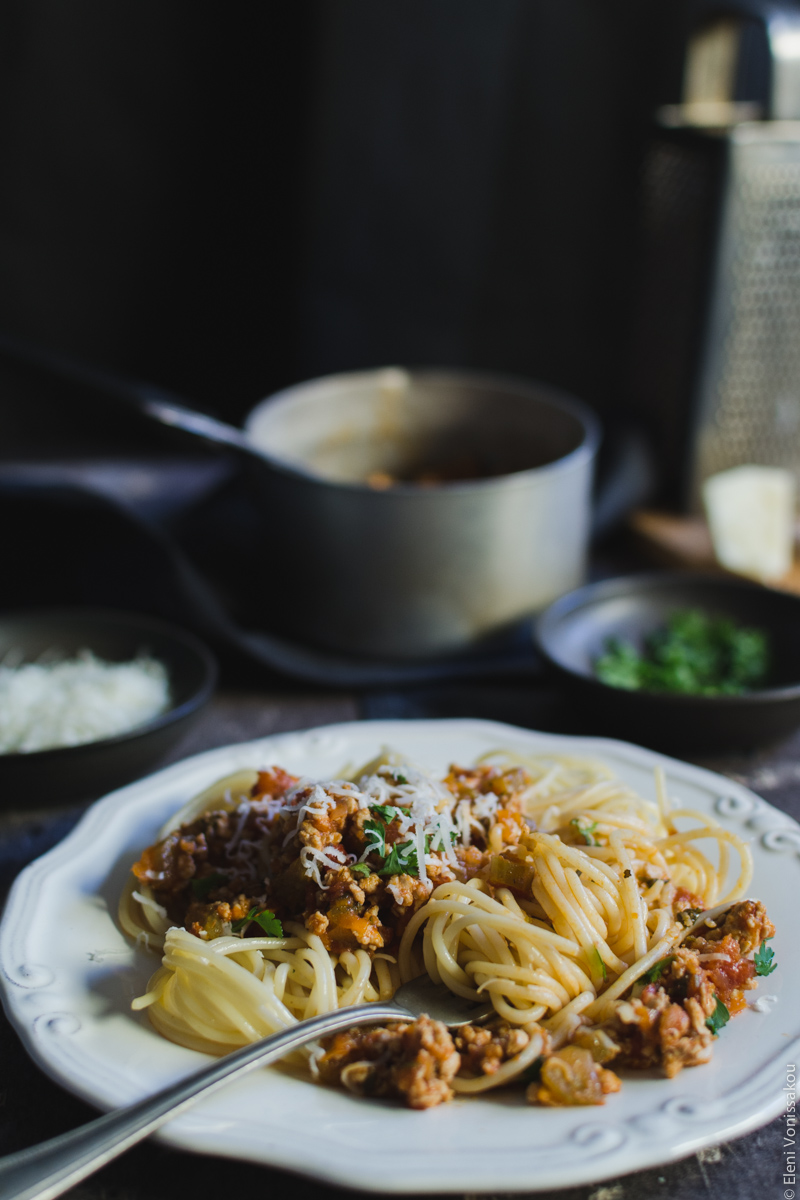 Slow Cooker Turkey Meat Sauce for Pasta (from scratch) www.thefoodiecorner.gr Photo description: A side view of a plate of spaghetti with sauce mixed in. A fork is sitting on the plate with some spaghetti wrapped around its tongs. In the background a small saucepan, a bowl of chopped parsley and a cheese grater (barely visible in the corner).