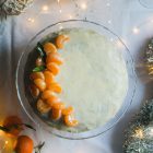 Greek New Year’s Cake (Vassilopita) with Mandarin, Almonds and Cardamom www.thefoodiecorner.gr Photo description: Top view of a cake covered in sugar glaze sitting on a glass cake stand in the middle of the photo. On the left side of the cake are some mandarin segments for decoration arranged in a half moon shape. To the top left and bottom right of the photo are some silver Christmas decorations (tinsel and baubles made of straw). To the bottom left are some whole mandarins with leaves attached. Everything is on a white tablecloth and there are tiny lights around the cake stand.