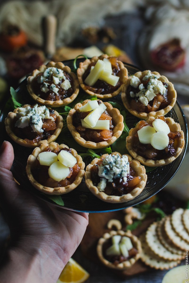 Savoury Tarts with Pear Chutney and Blue Cheese or Gruyere www.thefoodiecorner.gr Photo description: A hand entering the frame from the left side, holding a plate of tarts. Barely visible in the background is the rest of the cheese board.