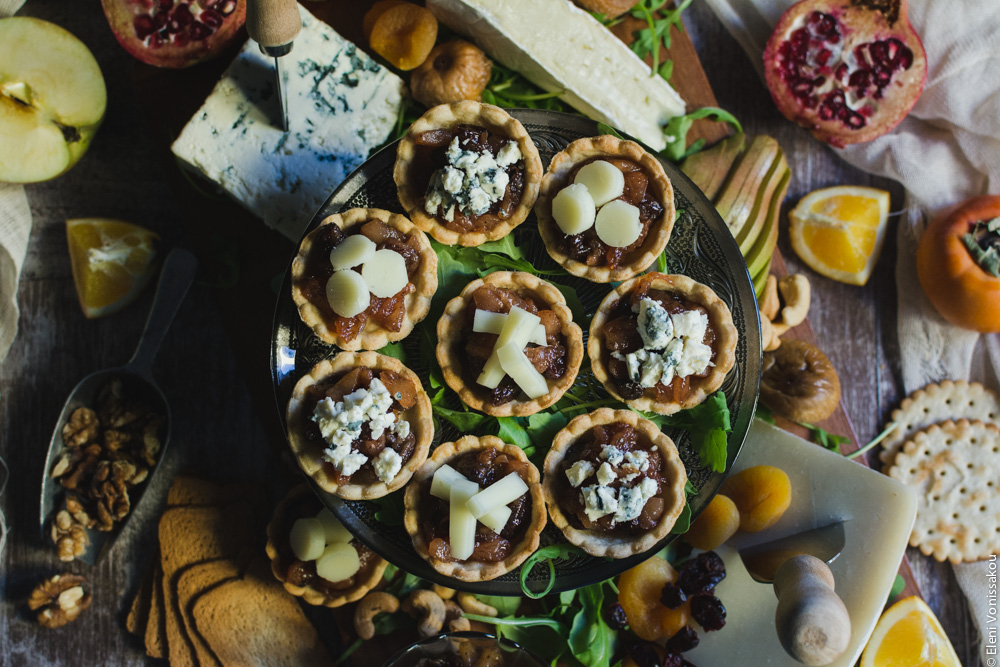 Savoury Tarts with Pear Chutney and Blue Cheese or Gruyere www.thefoodiecorner.gr Photo description: a top view of the tarts laid out on the cake stand in the middle of the cheese board. Around it are crackers, pieces of orange, pomegranate, gruyere cheese, blue cheese, brie, dried apricots and figs, walnut pieces, half an apple and a persimmon barely visible.