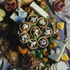 Savoury Tarts with Pear Chutney and Blue Cheese or Gruyere www.thefoodiecorner.gr Photo description: A wooden serving board filled with various cheeses, dried fruits, oranges, pears, persimmons, a cut pomegranate, nuts and crackers. In the centre is a cake stand with some savoury tarts on it. The edges of the tarts are fluted, they are filled with chutney and on top there are some pieces of gruyere or crumbled blue cheese. Barely visible under all the nibbles are some rocket (arugula) leaves.