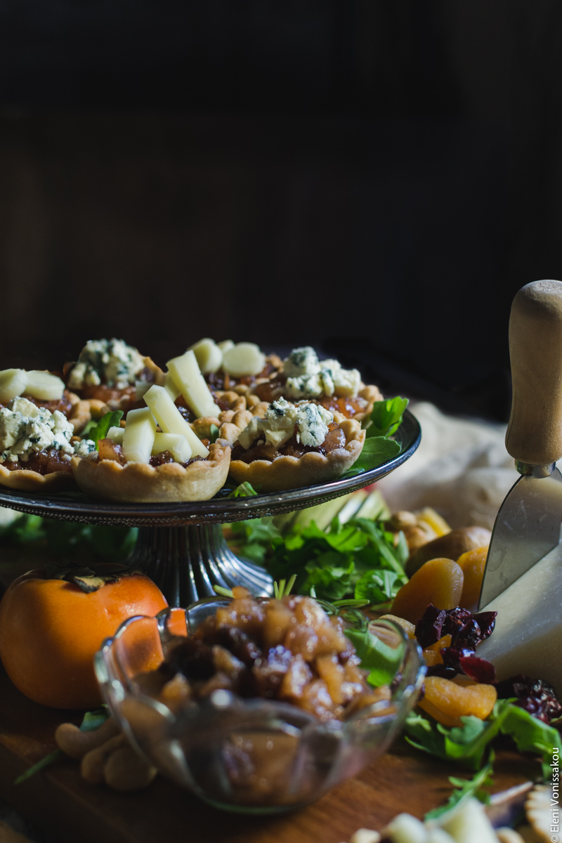 Savoury Tarts with Pear Chutney and Blue Cheese or Gruyere www.thefoodiecorner.gr Photo description: A side view of the cake stand with the tarts sitting on top. In the foreground is a small bowl of chutney and a persimmon. To the right, a piece of cheese with a cheese knife stuck in it. The background is dark.