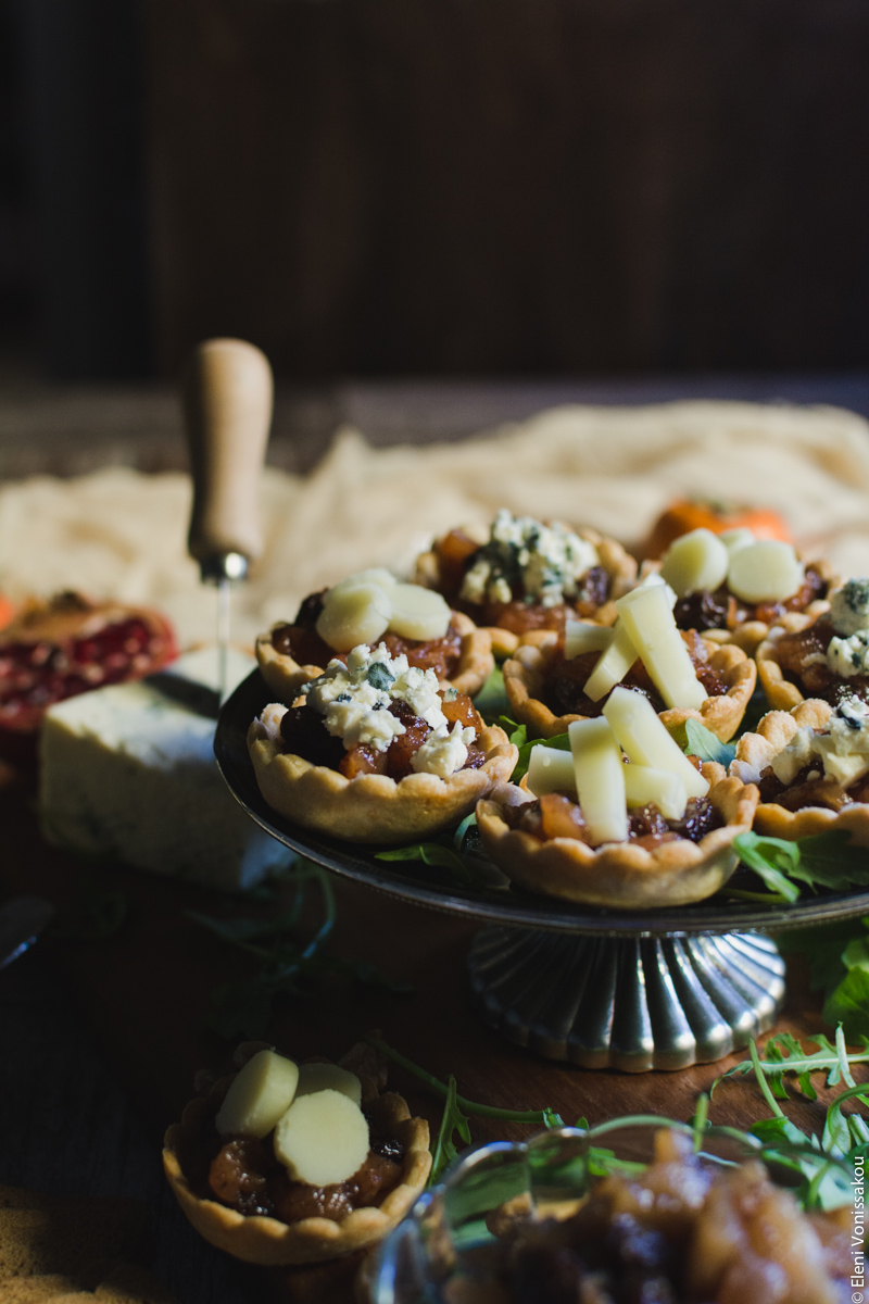 Savoury Tarts with Pear Chutney and Blue Cheese or Gruyere www.thefoodiecorner.gr Photo description: A side view of the tarts. In the foreground another tart on a wooden surface, the bowl of chutney barely visible and some rocket leaves scattered around. In the background a cheese knife sticking out of a piece of blue cheese and some muslin fabric further behind that. 