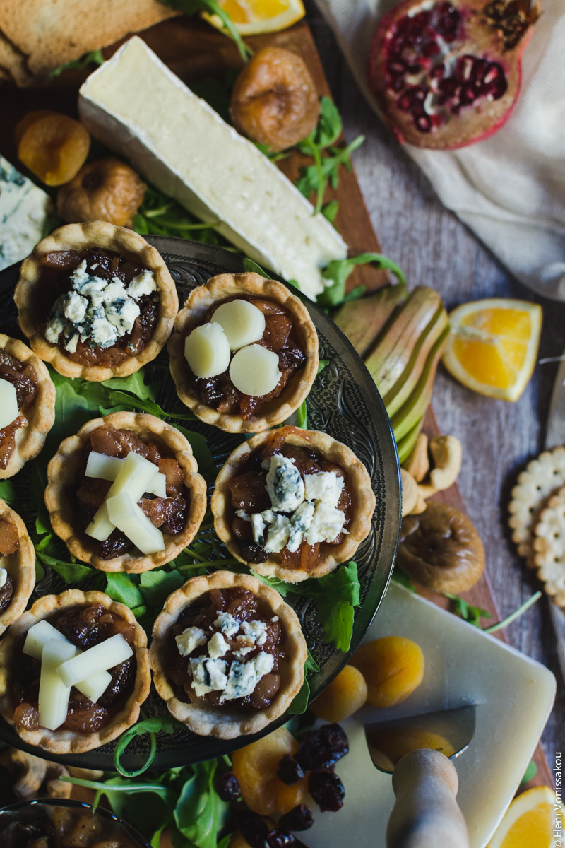 Savoury Tarts with Pear Chutney and Blue Cheese or Gruyere www.thefoodiecorner.gr Photo description: a closer top view of the tarts, some of them cut out of the photo on the left. In the background to the bottom of the photo is a piece of gruyere with a knife in it, and to the top, a piece of brie. Some fruit and crackers are also scattered around the tarts.