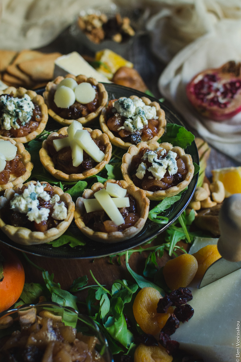 Savoury Tarts with Pear Chutney and Blue Cheese or Gruyere www.thefoodiecorner.gr Photo description: A three quarter view of the cake stand with the tarts sitting on top. In the foreground is a small bowl of chutney and some rocket leaves. To the right, a piece of cheese with a cheese knife stuck in it and some dried apricots. Towards the back, half a pomegranate on a piece of muslin fabric.