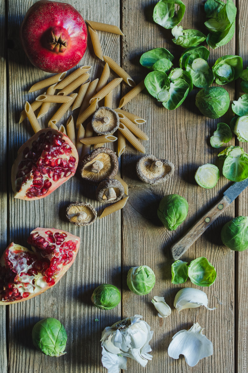 Pasta with Balsamic Roasted Brussels Sprouts and Shiitake Mushrooms (Vegan) www.thefoodiecorner.gr Photo description: To the left of the photo some broken pieces of pomegranate, and a whole one at the top. Scattered amongst them are some pieces of dried pasta and some dried mushrooms. To the right are Brussels sprouts leaves and a small knife. To the bottom of the picture a broken head of garlic.