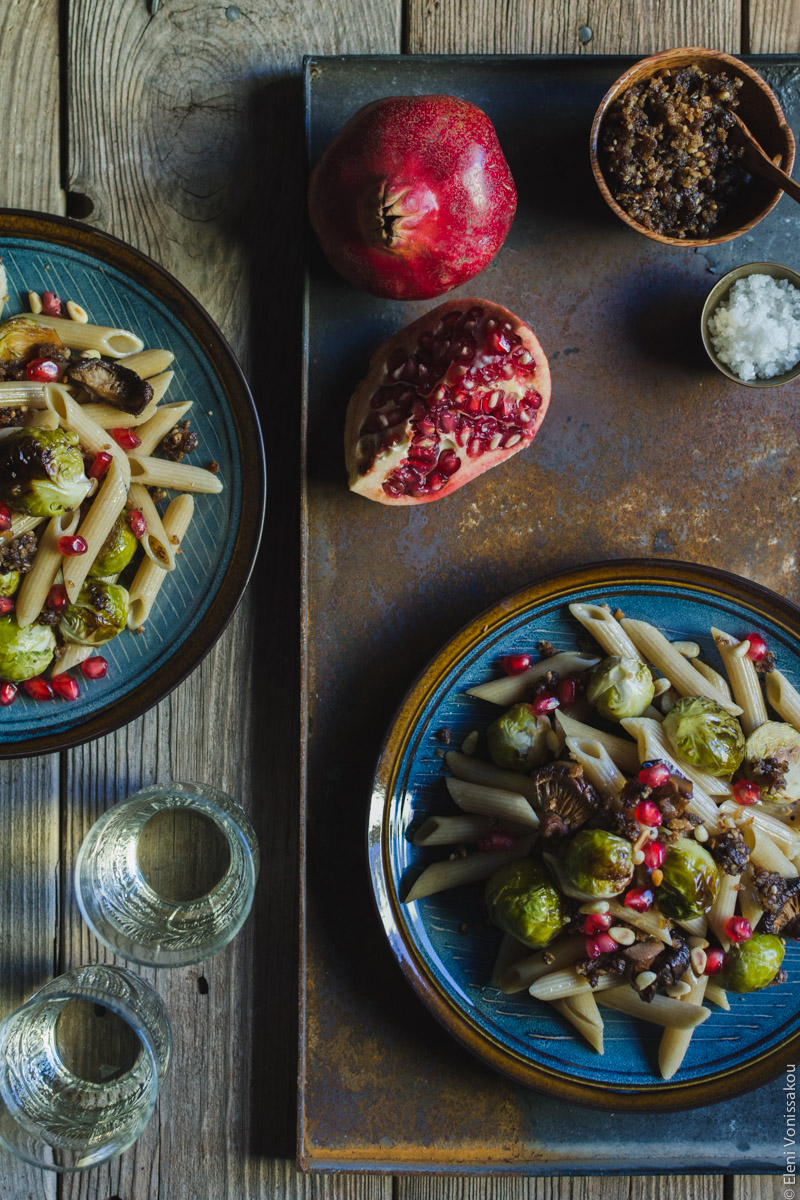 Pasta with Balsamic Roasted Brussels Sprouts and Shiitake Mushrooms (Vegan) www.thefoodiecorner.gr Photo description: Two plates of pasta, the one on the left half visible. The one on the right is on a rusty metal surface together with one whole pomegranate and a broken piece of another, a tiny bowl of salt and a wooden bowl of breadcrumbs. To the bottom left on the wooden surface are two glasses of wine.