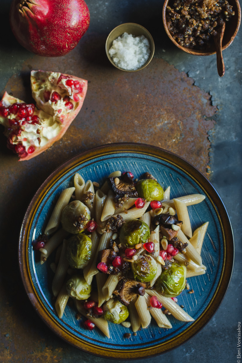 Pasta with Balsamic Roasted Brussels Sprouts and Shiitake Mushrooms (Vegan) www.thefoodiecorner.gr Photo description: One plate of pasta on a rusty metal surface. To the top, broken piece of pomegranate, a tiny bowl of salt and a wooden bowl with pangrattato.