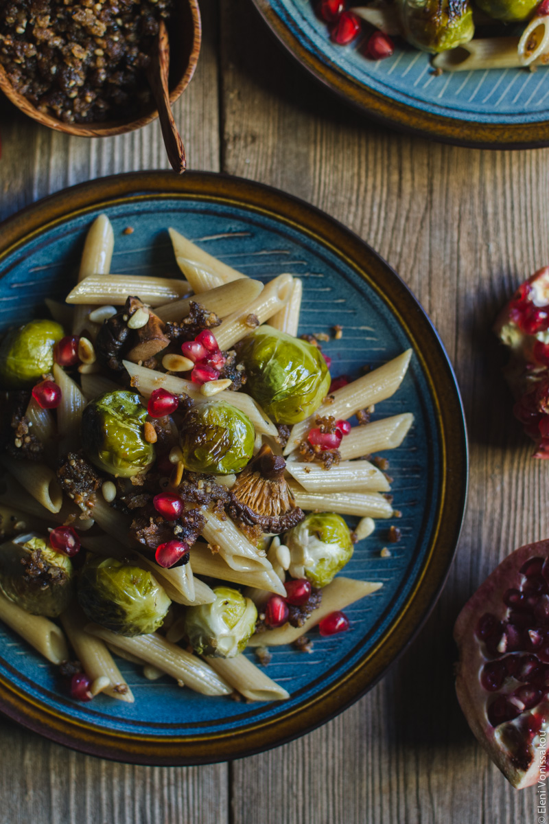 Pasta with Balsamic Roasted Brussels Sprouts and Shiitake Mushrooms (Vegan) www.thefoodiecorner.gr Photo description: A closer look at one of the plates of pasta. Among the penne are Brussels sprouts, pieces of mushroom and pomegranate arils. Barely visible to the top of the photo is the bowl with fried breadcrumbs.