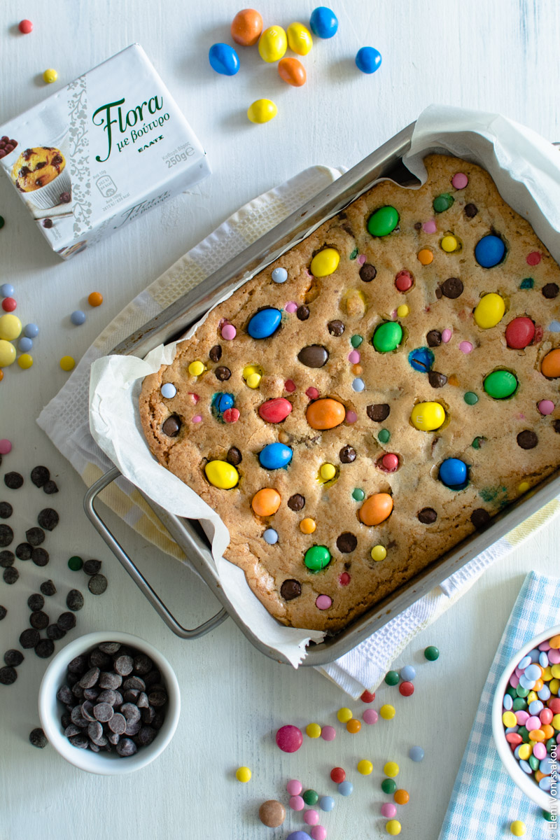 Εύκολα Cookie Bars με Κομμάτια Σοκολάτας και Πολύχρωμα Κουφέτα www.thefoodiecorner.gr Photo description: The cookie bars as they came out of the oven, before they were cut, still in the baking tin. The surface of the cookie is colourful with lots of candies on it. Around the tin are a block of margarine, some candies on the wooden surface and in a bowl, and some chocolate chips also on the surface and in a small bowl.