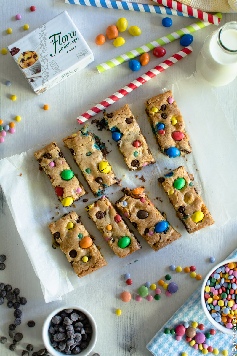 Εύκολα Cookie Bars με Κομμάτια Σοκολάτας και Πολύχρωμα Κουφέτα www.thefoodiecorner.gr Photo description: Eight cookie bars, arranged in two rows of four, on the baking paper they were baked on. Around them are the scattered candies and a block of margarine. In the top right corner is a small glass bottle of milk and some stripy paper straws.