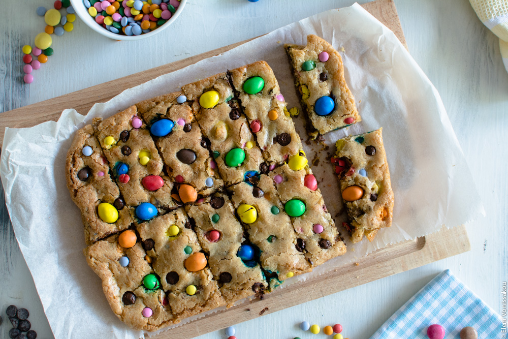 Easy, One-bowl, Colourful Chocolate Chip Cookie Bars www.thefoodiecorner.gr Photo description: A close view of the newly cut cookie bars, still arranged next to each other on the baking paper. The paper is set on a wooden chopping board. The top of the cookies is adorned with colourful chocolate candies.