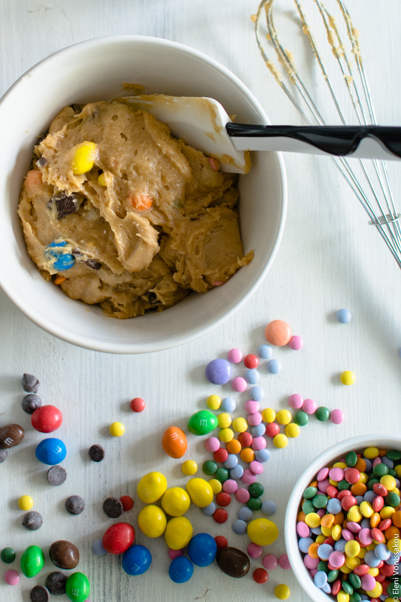 Easy, One-bowl, Colourful Chocolate Chip Cookie Bars www.thefoodiecorner.gr Photo description: An ingredient shot, with a mixing bowl at the top left of the photo, containing the cookie bar batter and a spatula. To the right of the bowl is a used whisk, and at the bottom of the photo are a small bowl with lots of chocolate candies (M&M’s, Smarties etc) with lots more scattered around.