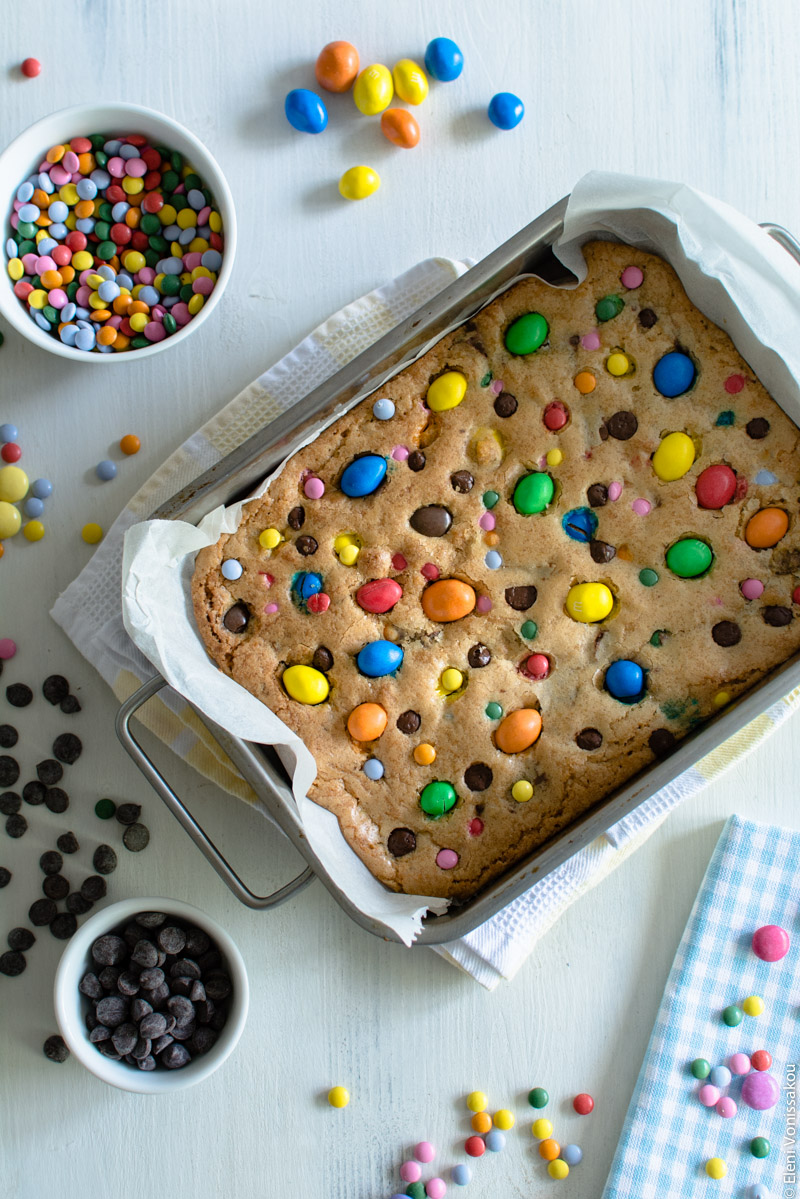 Easy, One-bowl, Colourful Chocolate Chip Cookie Bars www.thefoodiecorner.gr Photo description: The cookie bars as they came out of the oven, before they were cut, still in the baking tin. The surface of the cookie is colourful with lots of candies on it. Around the tin are some candies on the wooden surface and in a bowl, and some chocolate chips also on the surface and in a small bowl.