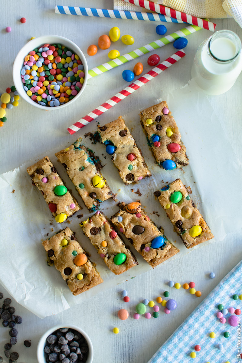 Easy, One-bowl, Colourful Chocolate Chip Cookie Bars www.thefoodiecorner.gr Photo description: Eight cookie bars, arranged in two rows of four, on the baking paper they were baked on. Around them are the scattered candies. In the top right corner is a small glass bottle of milk and some stripy paper straws.