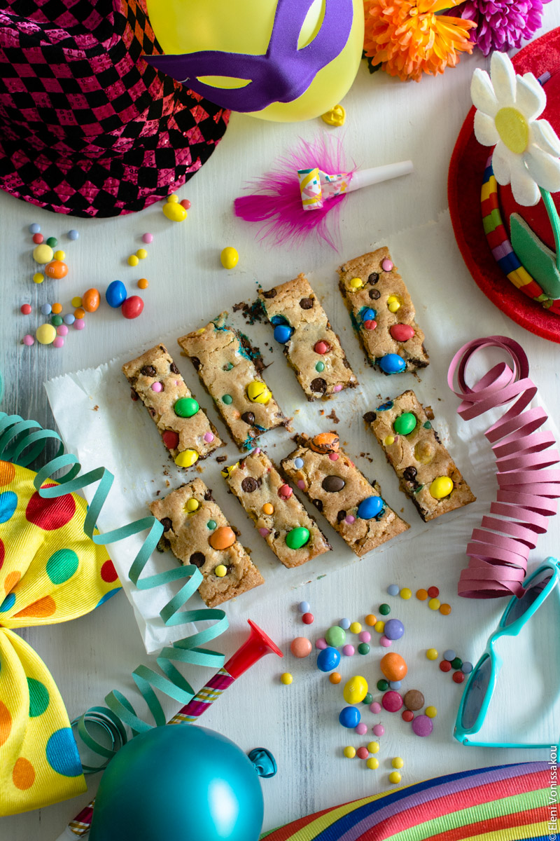 Easy, One-bowl, Colourful Chocolate Chip Cookie Bars www.thefoodiecorner.gr Photo description: The cookie bars are in the centre of the photo surrounded by lots of carnival items. Clockwise from top right, a clown hat with a flower sticking out of it, some paper streamers, a pair of plastic turquoise sunglasses, some stripey material (barely showing), a balloon, a big clown bow tie, a shiny hat, a party horn and a balloon with an eye mask on it.  Around the cookies are some scattered colourful candies too.