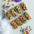 Εύκολα Cookie Bars με Κομμάτια Σοκολάτας και Πολύχρωμα Κουφέτα www.thefoodiecorner.gr Photo description: Cookie bars with colourful chocolate candies on top. The bars are on a white wooden surface, arranged in two rows of four. To their top left and bottom right are some more candies. Towards the top of the photo in the centre is a purple carnival eye mask, and the bottom left corner of the photo is filled with white feathers.