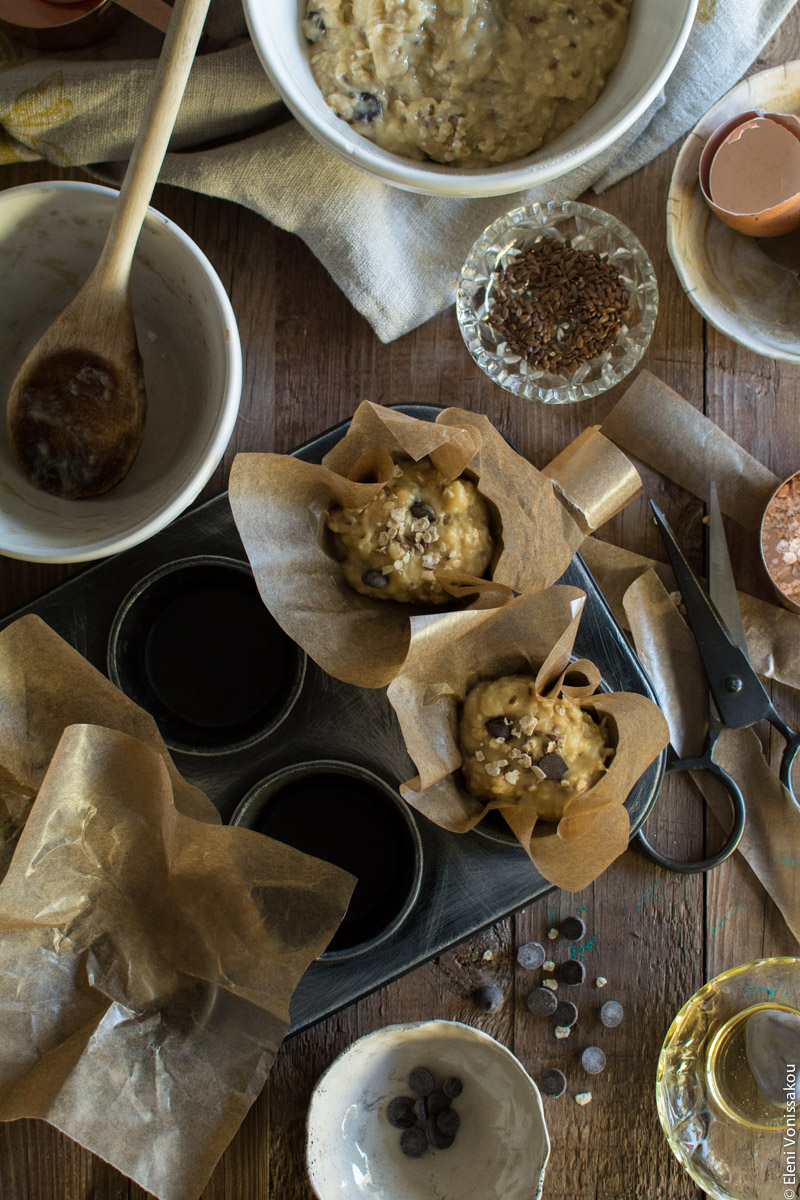 Healthy Banana Muffins with Oats, Tahini, Honey and Chocolate Chips www.thefoodiecorner.gr Photo description: A slightly battered and old muffin tin with two holes already filled with batter. The batter is sitting in square pieces of grease-proof paper which is serving as muffin liners. To the left are two more pieces of paper waiting to be filled. Clockwise from the top left are an empty bowl and a used wooden spoon, a bowl of batter (barely visible), a small glass dish with flax seeds, some cut up grease proof paper, a pair of metal scissors, an empty bowl with the remains of honey and a bowl with a few chocolate chips. Some chips are also scattered on the wooden surface.