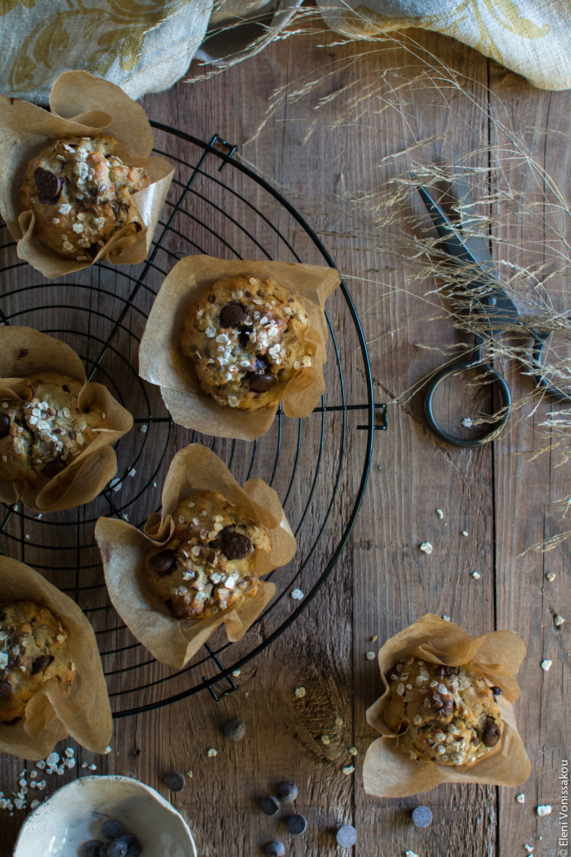 Healthy Banana Muffins with Oats, Tahini, Honey and Chocolate Chips www.thefoodiecorner.gr Photo description: A round wire rack half visible on the left side of the photo, with some of the muffins on top. To the right the metal scissors and to the bottom right another muffin and some scattered chocolate chips. 