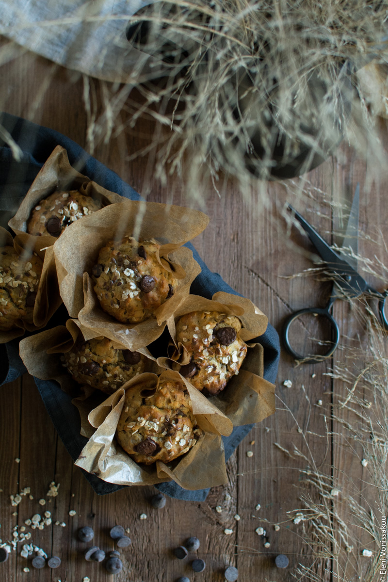 Healthy Banana Muffins with Oats, Tahini, Honey and Chocolate Chips www.thefoodiecorner.gr Photo description: Top view of the muffins inside a rectangular baking tin lined with a linen napkin. The tin is half visible towards the left of the photo. At the top of the photo are some dried grass leaves, hanging over the muffins partly obscuring the view. To the bottom right of the photo are some more grass leaves lying on the wooden surface. Towards the bottom left are some scattered oat flakes and chocolate chips.