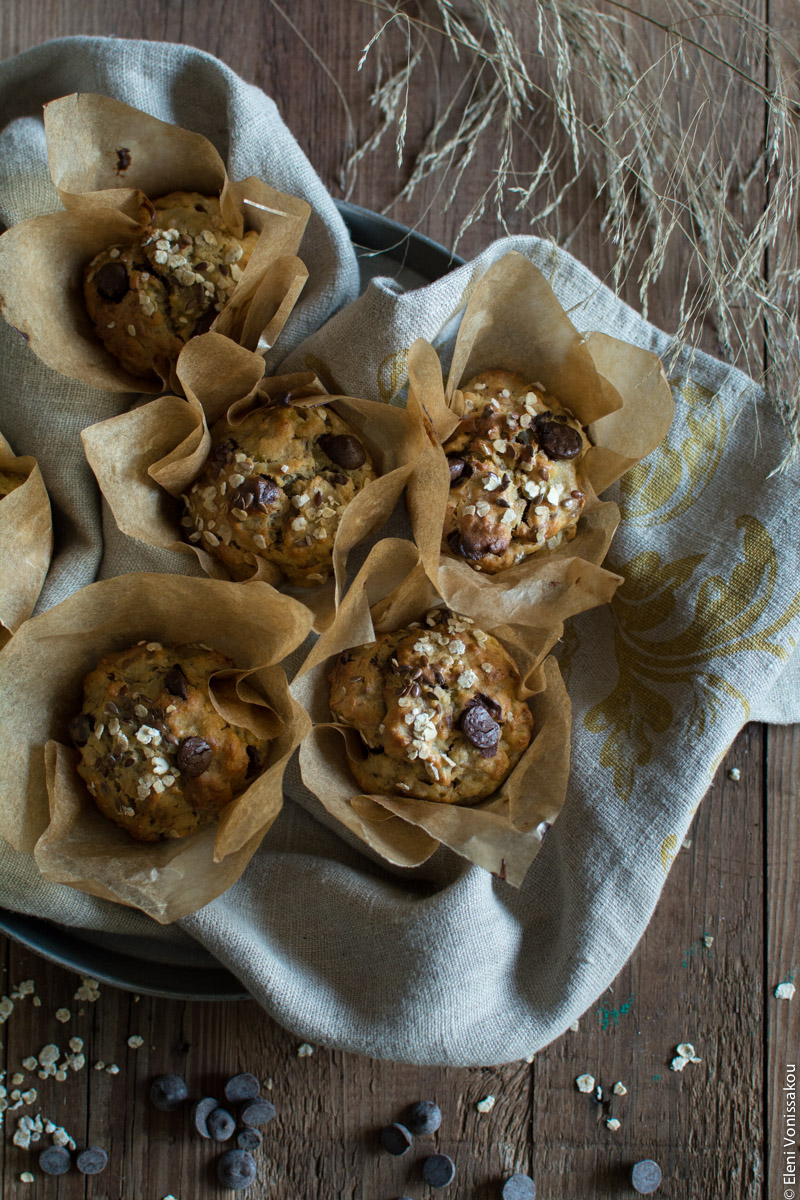 Healthy Banana Muffins with Oats, Tahini, Honey and Chocolate Chips www.thefoodiecorner.gr Photo description: Top view of a dish lined with a linen tea towel, filled with muffins. To the top of the photo are some dried grass leaves lying on the wooden surface and to the bottom are some scattered oat flakes and chocolate chips.