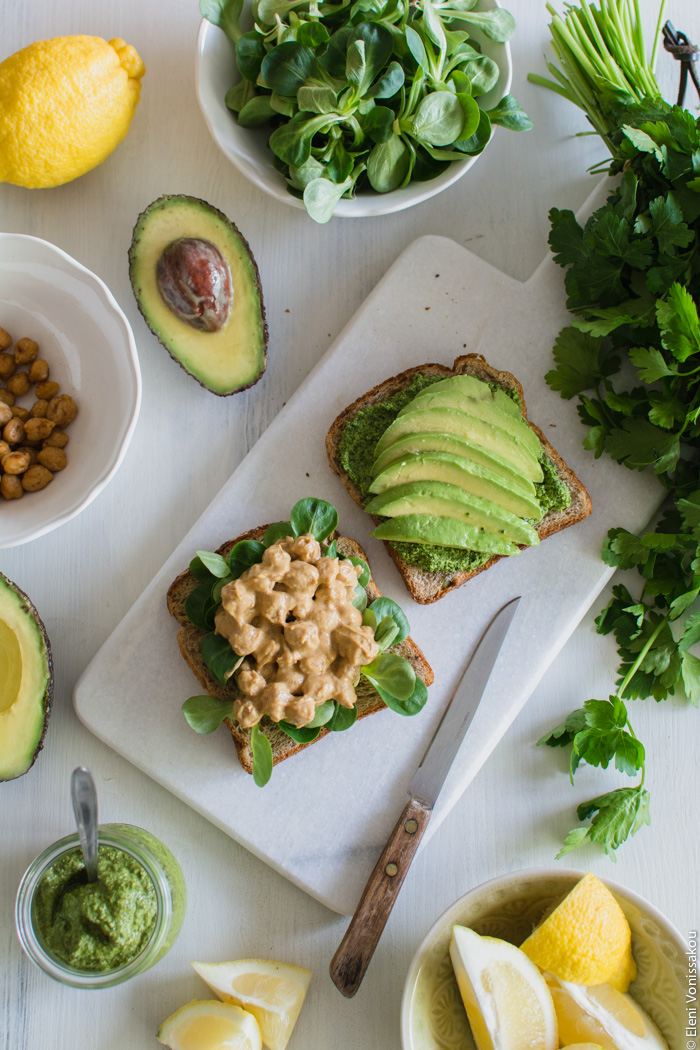 Vegan Chickpea and Tahini Sandwich with Avocado and Parsley Pesto www.thefoodiecorner.gr Photo description: Two slices of bread, one of them spread with parsley pesto and slices of avocado, the other with lamb’s lettuce and chickpeas in tahini sauce. Both slices are on a marble cutting board surrounded by more ingredients.