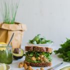 Vegan Chickpea and Tahini Sandwich with Avocado and Parsley Pesto www.thefoodiecorner.gr Photo description: A front side view of a sandwich, stuffed with filling. The sandwich is on a marble cutting board, which in turn is on a white wooden surface. It is surrounded by parsley, avocado, some lemon wedges, a small jar of pesto and a pot of chives in the background.