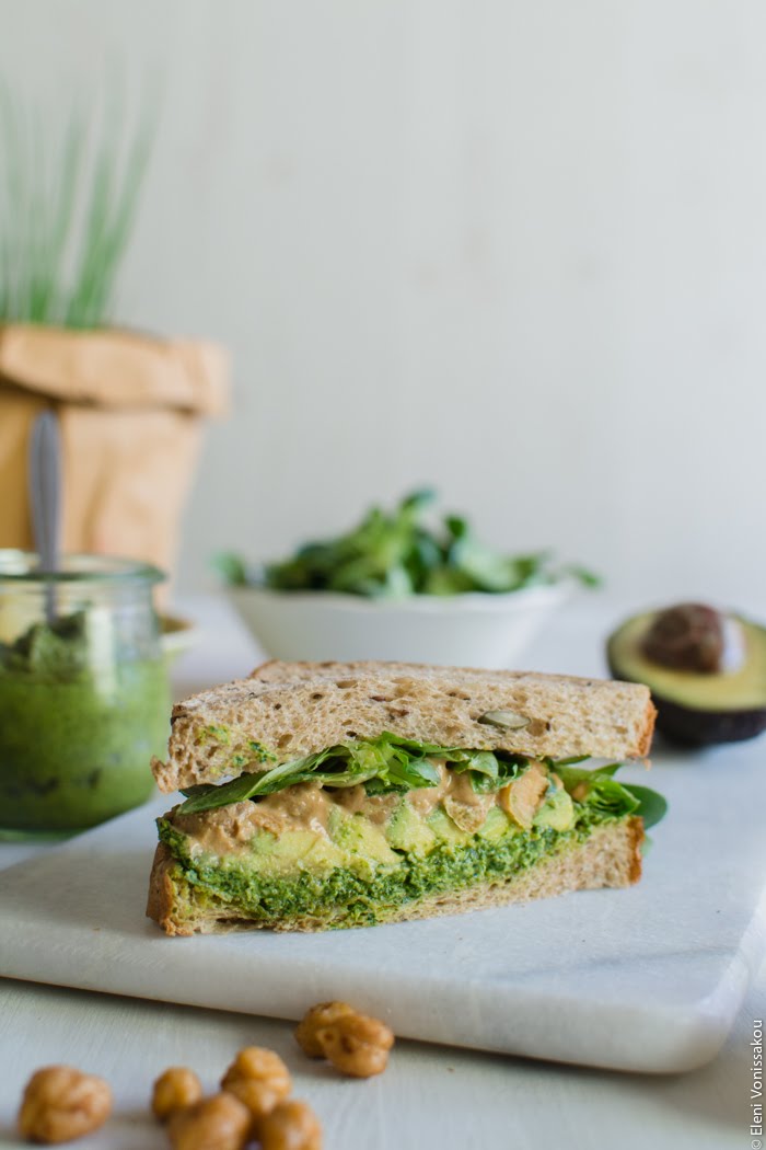 Vegan Σάντουιτς με Αβοκάντο, Πέστο Μαϊντανού και Ρεβίθια με Σος από Ταχίνι και Πετιμέζι www.thefoodiecorner.gr Photo description: A side view of a cut sandwich, the layers of ingredients clearly visible between the bread. In the background, a small jar of pesto, a pot of chives, and a bowl of lamb’s lettuce. 