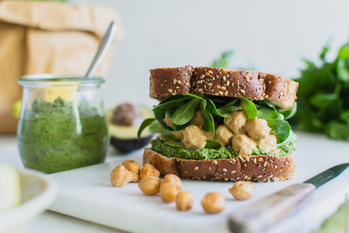 Vegan Chickpea and Tahini Sandwich with Avocado and Parsley Pesto www.thefoodiecorner.gr Photo description: A front side view of the sandwich, close up - the ingredients spilling out from between the bread. To the left, the jar of pesto with a spoon in it.