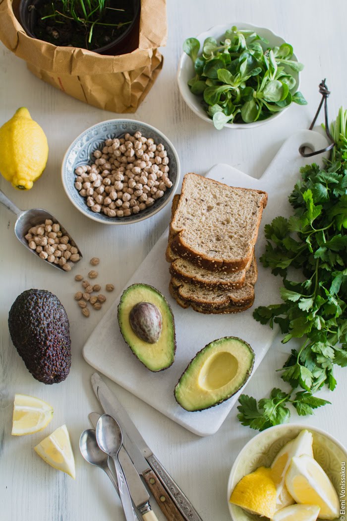 Vegan Σάντουιτς με Αβοκάντο, Πέστο Μαϊντανού και Ρεβίθια με Σος από Ταχίνι και Πετιμέζι www.thefoodiecorner.gr Photo description: A top view of the ingredients laid out. Clockwise from top right, a bowl of lamb’s lettuce, a bunch of parsley, some slices of whole-wheat bread, a halved avocado, a bowl of lemon wedges, some cutlery, a whole avocado, some dried chickpeas in a bowl and in a tiny spice shovel, and a flower pot with chives in the background.