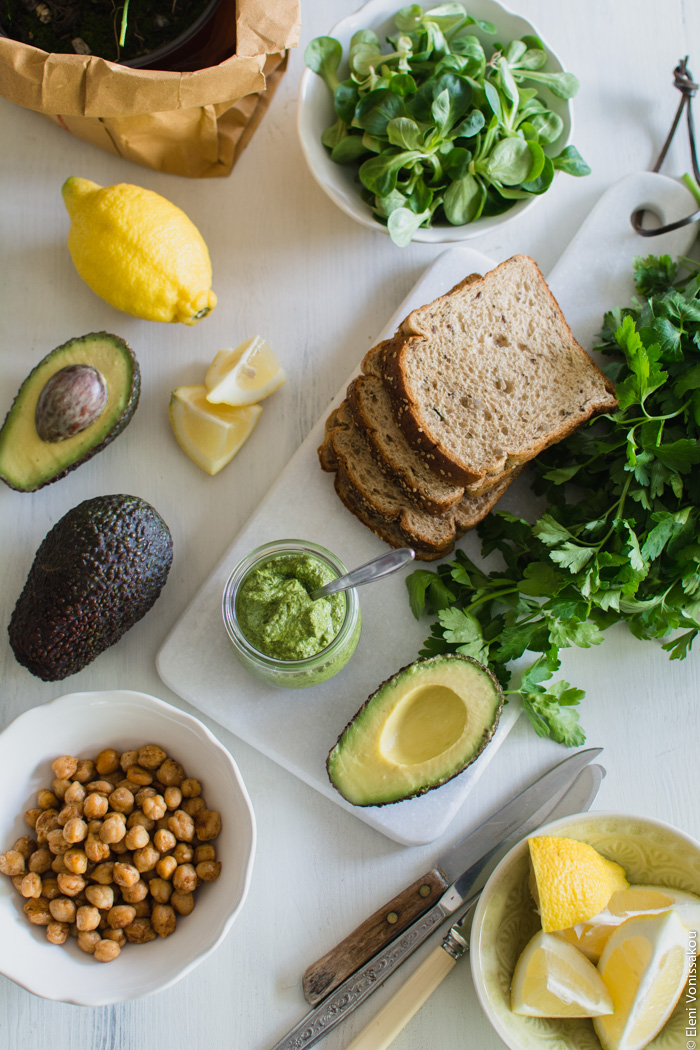 Vegan Chickpea and Tahini Sandwich with Avocado and Parsley Pesto www.thefoodiecorner.gr Photo description: A top view of the ingredients. Sliced whole-wheat bread, a bunch of parsley, one whole and one halved avocado, a whole lemon and a bowl of lemon wedges, a small jar of pesto, a bowl of sautéed chickpeas, and a bowl of lamb’s lettuce.