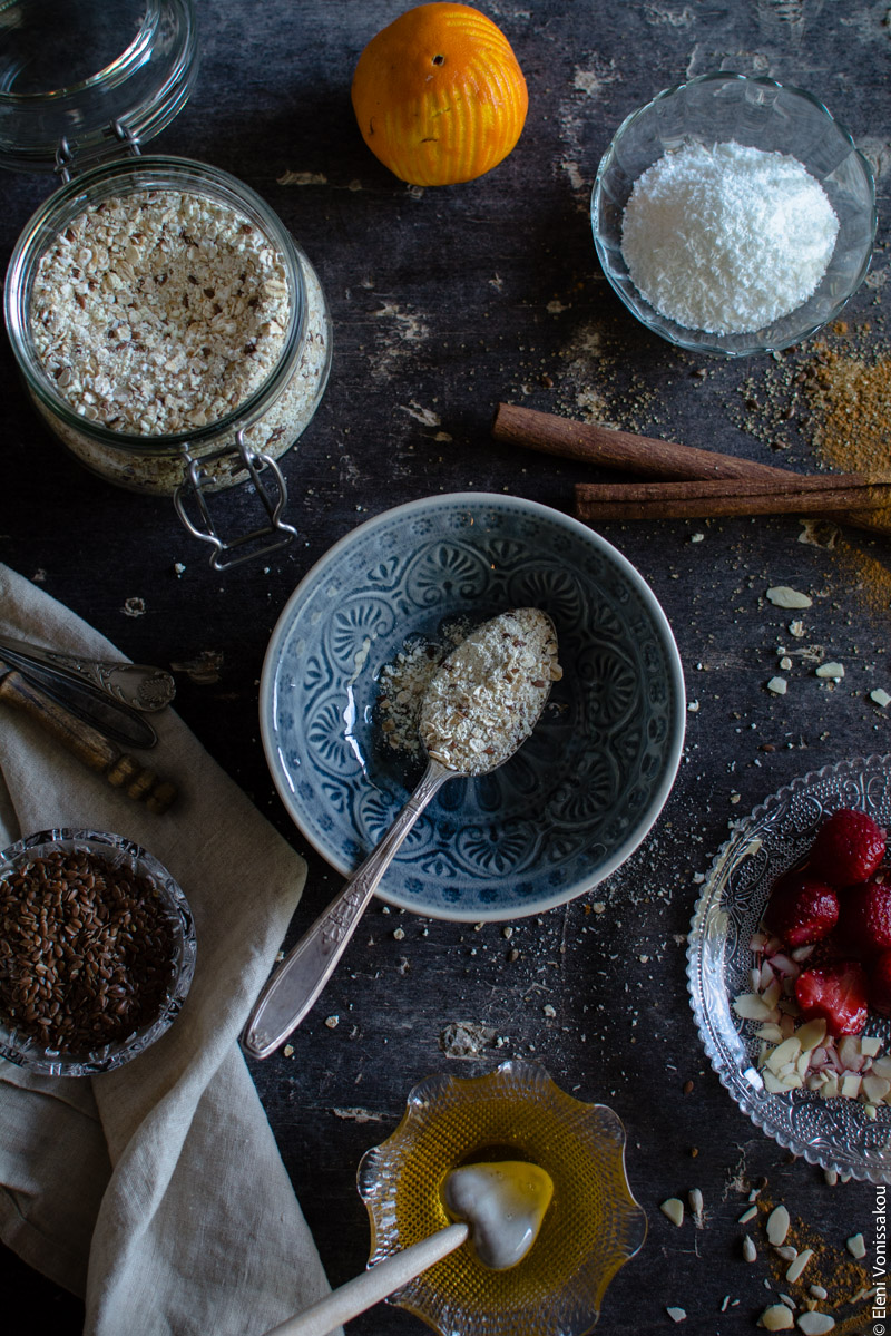 Oat Mix for Instant, Creamy Microwave Porridge (Oatmeal) www.thefoodiecorner.gr Photo description: An empty bowl with a spoon inside. The spoon has oat mix on it. Around the bowl are various other jars and bowls of ingredients. Clockwise from top, a zested orange, a small bowl of coconut flakes, two sticks of cinnamon, a glass dish of strawberries and almond flakes, a small bowl of honey with a honey stick inside, a linen napkin with a tiny bowl of flaxseed sitting on it, and a mason jar with the oat mixture.