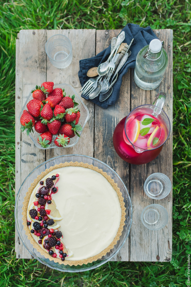 Lemon Curd and Mascarpone Cheesecake with Berries www.thefoodiecorner.gr Photo description: An overhead view of the box on the grass. On the box are the cheesecake, a bowl of strawberries, a jug of pink lemonade, some empty glasses, a bottle of water and a linen napkin with cutlery on it.