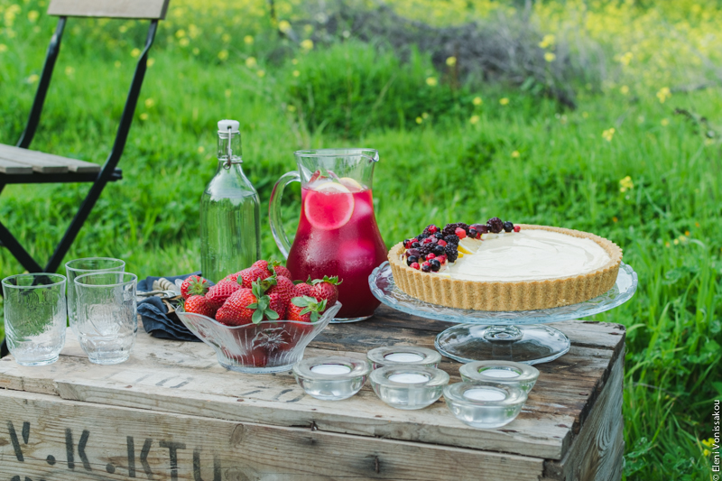 Lemon Curd and Mascarpone Cheesecake with Berries www.thefoodiecorner.gr Photo description: A ¾ view of the box with the cheesecake, the jug of lemonade and the bowl of strawberries. To the left, part of a chair.